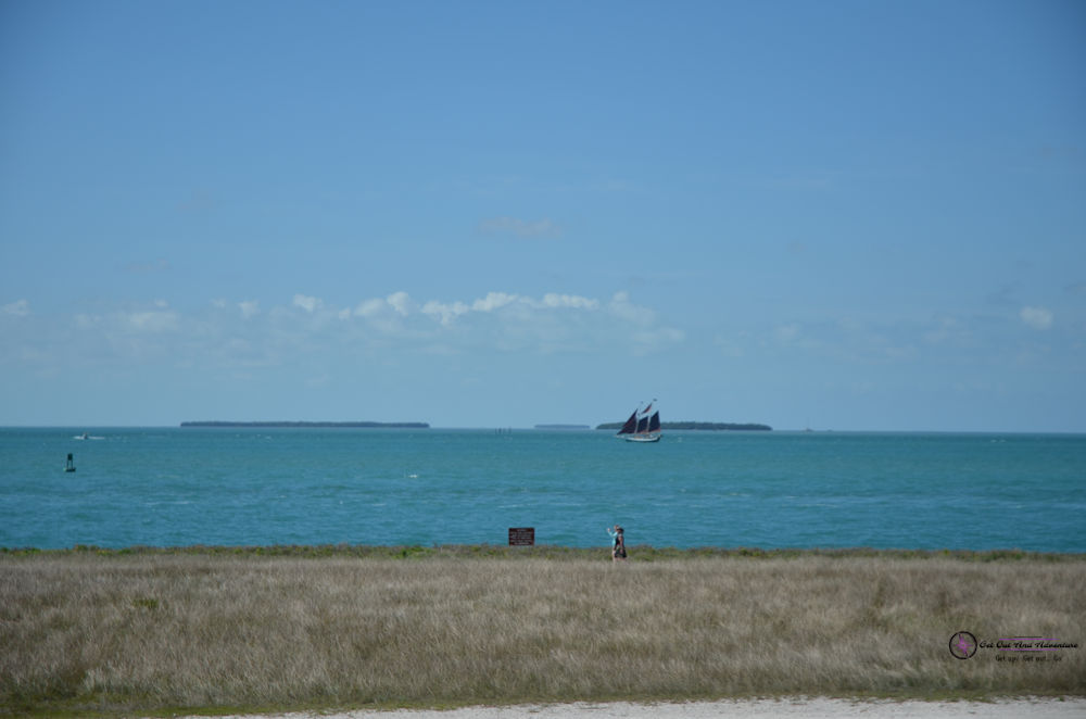 A three masted sailboat sits in the waters outside of Fort Zachery Taylor State Park under powder blue skies.