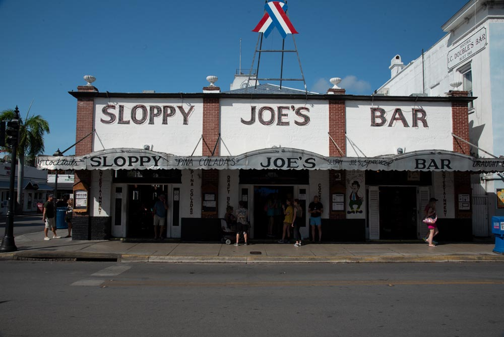 Sloppy Joes Bar on Duval Street. The Brick columns are separated by white stucco. A grey overhang shades the sidewalk in front with the words "Sloppy Joes Bar"