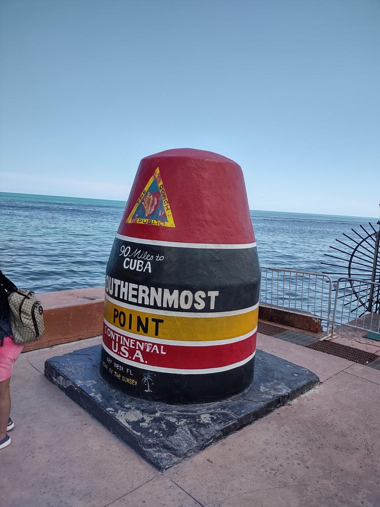 The red, black, and yellow buoy that marks the southernmost point of the USA sits in front on the turquoise waters of the Atlantic Ocean. Welcoming visitors on your Key West Vacation.