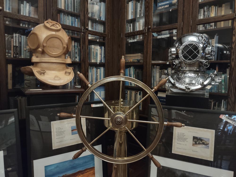 An old ship's steering wheel sits in between two old dive helmets in front of glass encased bookshelves in the History of Diving Museum library.