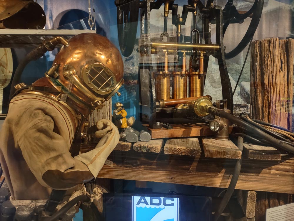 A figure in an old brown canvas and rubber dive suit with a brass helmet looks over a tool bench of diving equipment at the History of Diving Museum 