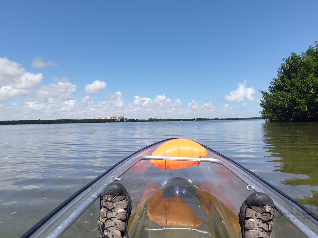 Looking over a clear kayak, you can see calm water and mangrove trees lining the shore with a building peaking above the tree line.