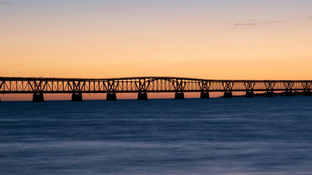The sunset gives the sky behind the iconic Bahia Honda Bridge an orange glow contrasting with the blue waters at Bahia Honda State Park, a must visit for your first time visiting the Florida Keys.