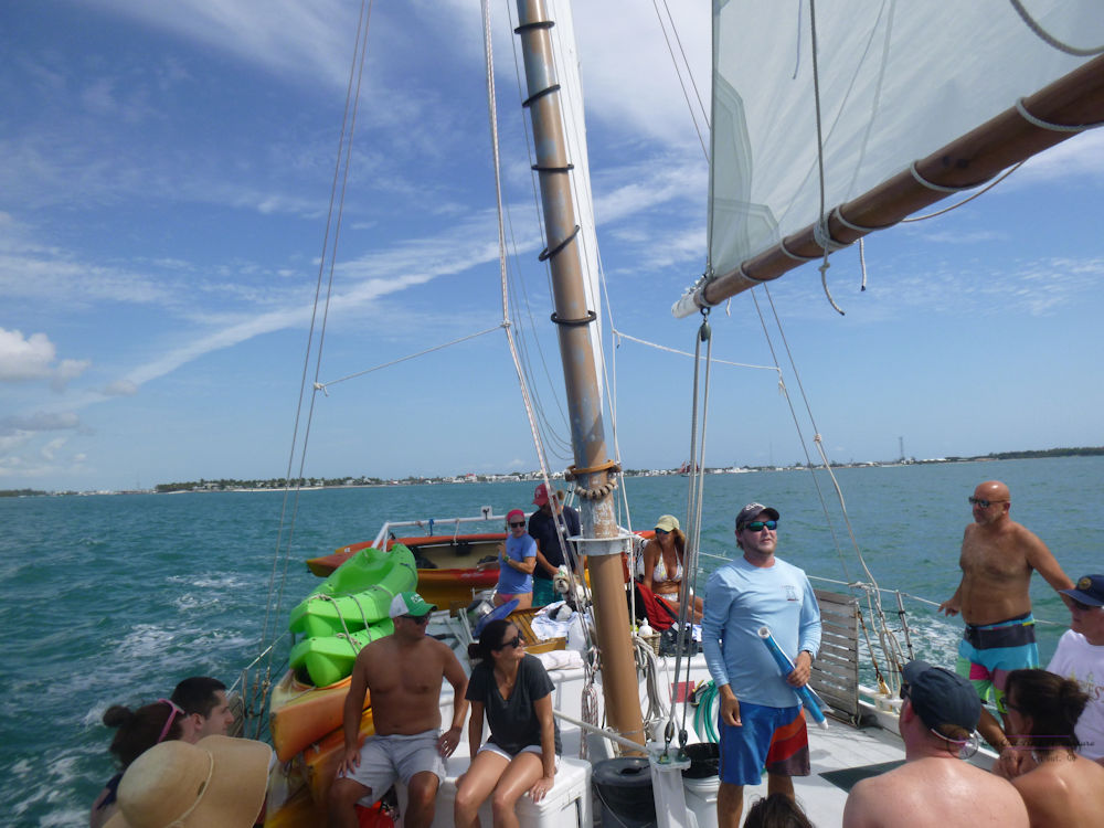 A gathering of people on a sailboat deck with Key West in the background surrounded by turquoise water.