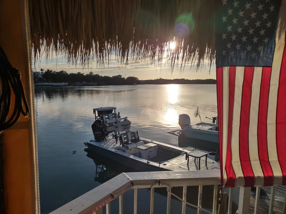 A USA flag hangs off of a tiki hut while looking across a bay with the sunsetting in the background