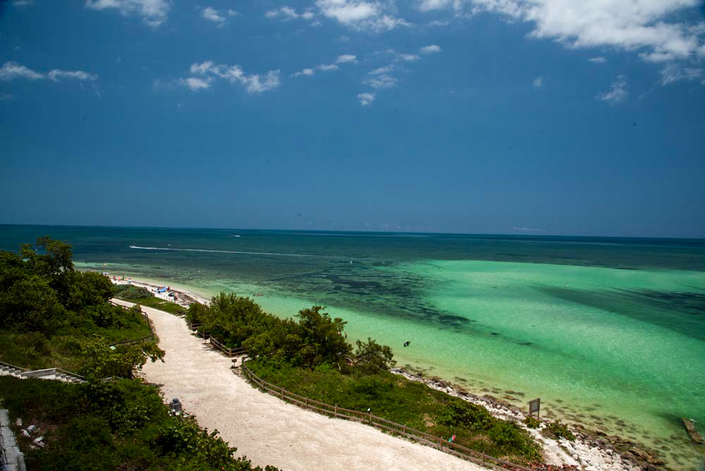 A winding gravel path sits on the edge of a water in Bahia Honda State Park.  The clear water shows the tan sand and sea grass further out.  Eventually the water turns blue under blue skies with scattered clouds.  Bahia Honda should be on your Florida Keys Checklist.