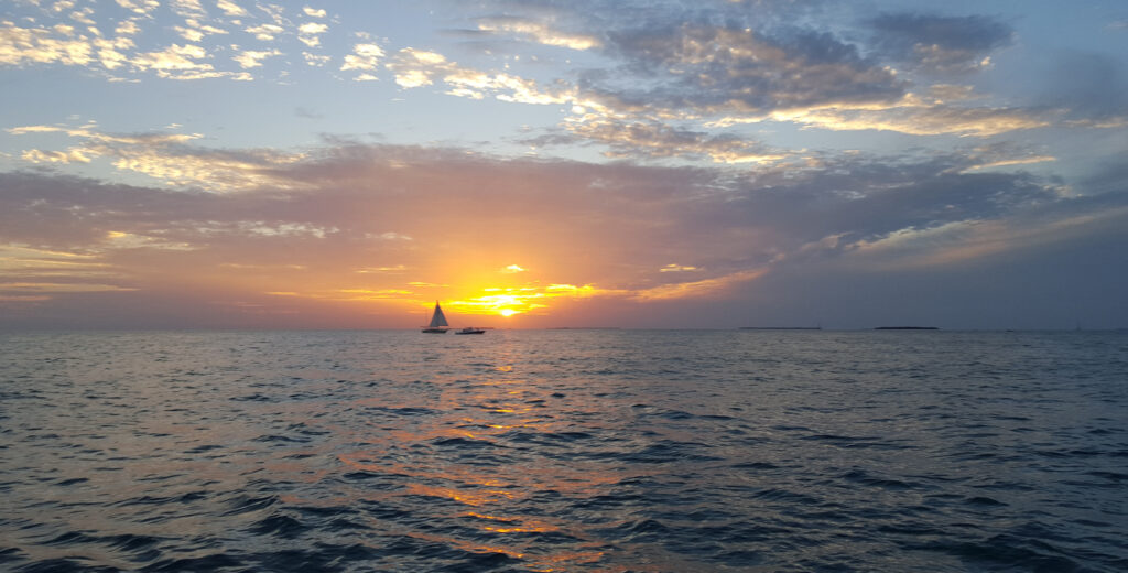 A sailboat and a powerboat cut across the deep blue seas of the Atlantic Ocean in Key West.  In the background the sun is just above the water shining yellow and orange.