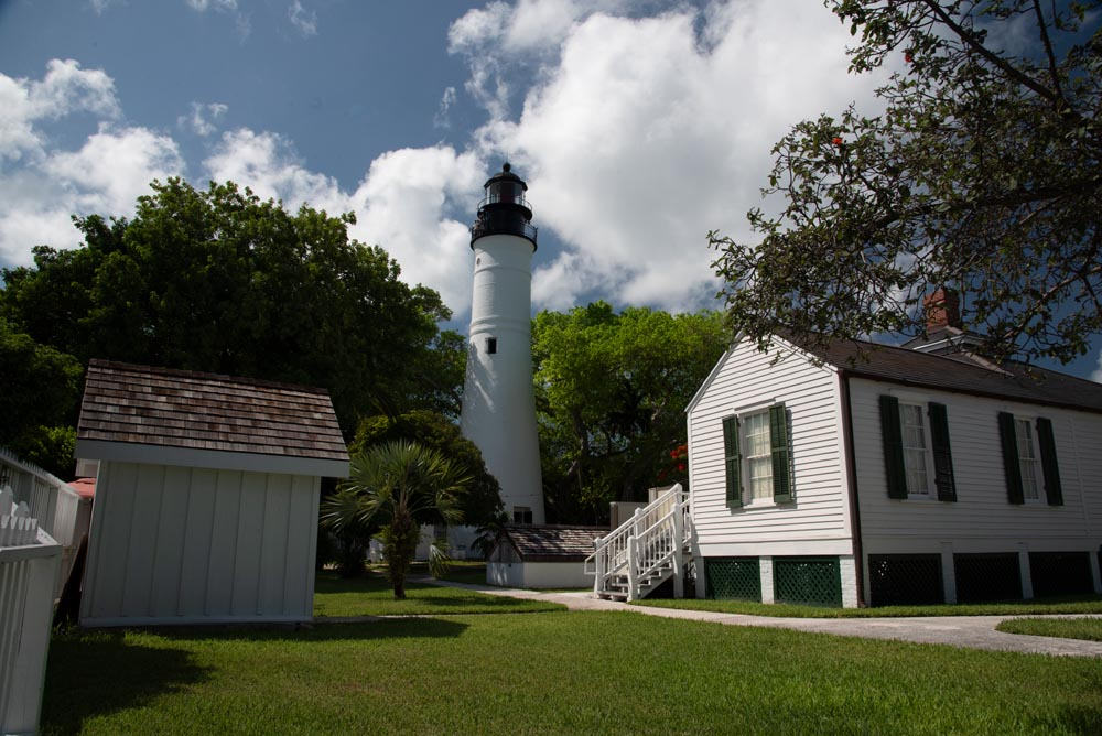 The white lighthouse of Key West towers over the green grass and white clapboard keepers' quarters. White clouds hang lazily in the blue sky.
